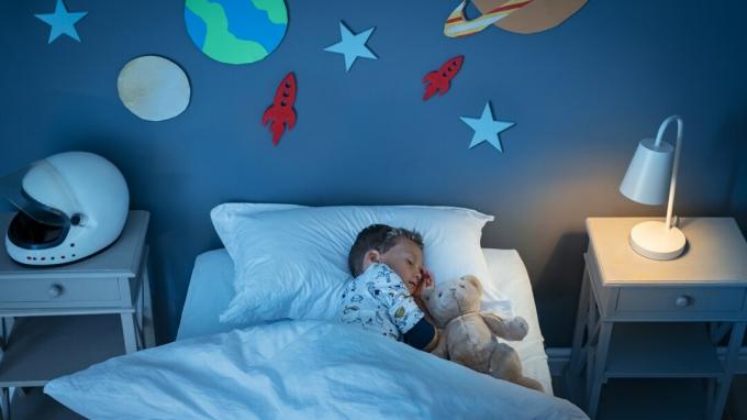 Поглед из високог угла на малог детета из снова који сања да постане астронаут док спава с медведом у соби уређеној свемиром.